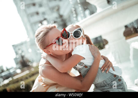 Liebevolle Familie. Gerne schöne Frau umarmte ihre kleine Tochter lächelnd vor dem weißen Brunnen außerhalb. Stockfoto