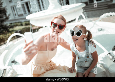 Eine gute Zeit. Fröhliche Mutter und Tochter in der Nähe von der schönen Brunnen sitzen und lustige Gesichter unter selfie. Stockfoto