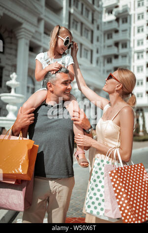 Darauf achten. Lächelnd schöne Frau neuanordnen Sonnenbrille ihrer Tochter sitzen auf den Schultern ihres Vaters nach dem Einkaufen. Stockfoto