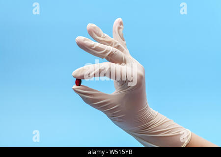 Nahaufnahme einer Hand Fragment in einer weißen medizinischen Handschuhen Holding eine Tablette, Kapsel auf blauem Hintergrund. Medizinischer Hintergrund Stockfoto