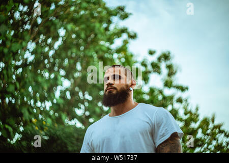 Porträt der jungen bärtigen Mann stand draußen vor der grünen Bäume und der blaue Himmel im Hintergrund Stockfoto