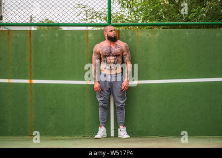 Junger Mann mit Tattoos und Bart trug Trainerhosen und kein Hemd lehnte sich auf Tennisplatz an der Wand und auf Seite Stockfoto
