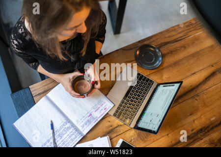 Hübsche junge Frau am Tisch mit Notebook, Planer, Smartphone und Laptop im Cafe Holding heiße Schokolade Tasse Stockfoto