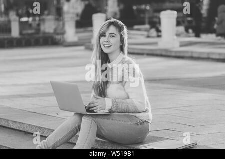 Hübsche junge fröhliche Teenager Frau studieren oder arbeiten am Laptop sitzen glücklich und auf Treppen in der Stadt draußen im städtischen Hintergrund. In der studentischen Stockfoto