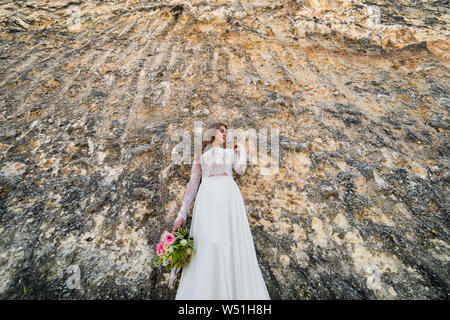 Portrait von hübschen Braut im weißen Kleid mit Blumen und lehnte sich auf Felswand auf der Suche nach Side, Low Angle Shot Stockfoto