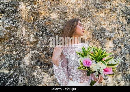 Schöne Braut im weißen Kleid mit Blumen in der Hand und die Haare Berühren mit anderen, lehnte sich gegen die Felswand in Bali. Stockfoto
