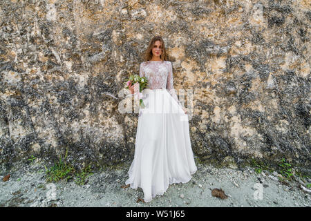 Porträt der schönen blonden Braut im weißen Kleid mit Blumen in der Nähe der Felswand Stockfoto