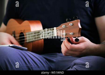 Ein Kerl in einem dunkelblauen t-shirt und lila Jeans sitzt mit dem Telefon auf seinem Schoß und passt der Sound der Ukulele hält er. Stockfoto