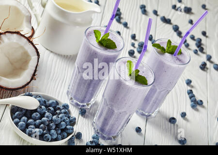 Blueberry schaumigen Milchshakes von Kokosmilch in drei Gläser mit Strohhalmen. Zutaten auf einem weißen Holztisch, horizontale Ansicht von oben Stockfoto