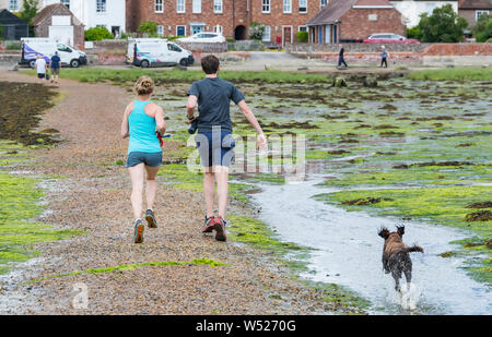 Ein junges Paar, das mit einem Hund über Bosham Causeway bei Ebbe in Chichester Harbour, Bosham Village, West Sussex, England, UK. Stockfoto