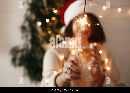 Frohes Neues Jahr Silvester Party Atmosphäre. Brennende wunderkerze in der Hand von stilvollen Mädchen in Santa Hut auf dem Hintergrund der modernen Weihnachtsbaum Licht in dunklen Zimmer. Stockfoto