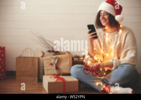 Verschwommenes Bild von stilvollen Happy girl in Santa hat im Bildschirm "Telefon" in festlichen Weihnachtsbeleuchtung auf Hintergrund o Geschenke und Geschenke in moderne Zimmer Stockfoto