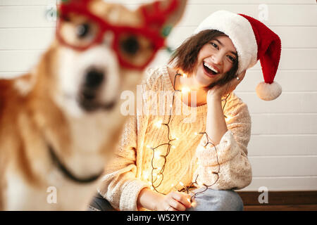 Stilvolle Mädchen lächelnd in Santa Hut und Weihnachtsbeleuchtung und niedlichen goldenen Hund mit lustigen Emotionen im festlichen Rentier Gläser mit Geweih. Mir Stockfoto