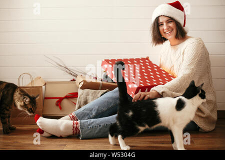 Stilvolle Happy girl in Santa Hut und gemütlichen Pullover Eröffnung Weihnachten Geschenk Box und Spielen mit Katzen in geschmückten Zimmer. Junge hipster Frau hol Stockfoto