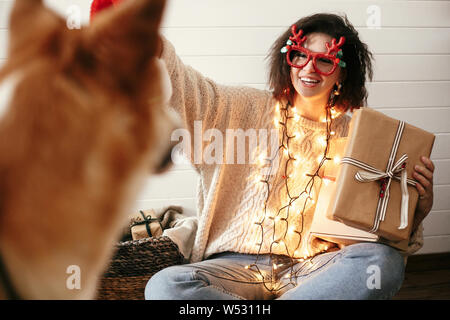 Stilvolle glückliches Mädchen mit Geschenk Box in Weihnachtsbeleuchtung und lächelnd cute goldenen Hund. Junge Frau im festlichen Gläser mit Hirschgeweihen und Stockfoto