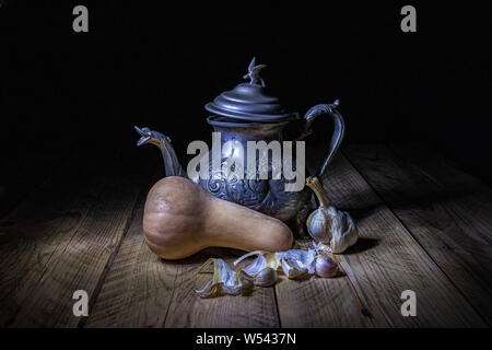 Noch immer leben gegen den dunklen Hintergrund. Osten Wasserkocher aus Metall auf einem Holztisch mit Kürbis und Knoblauch vor ihm. Stockfoto