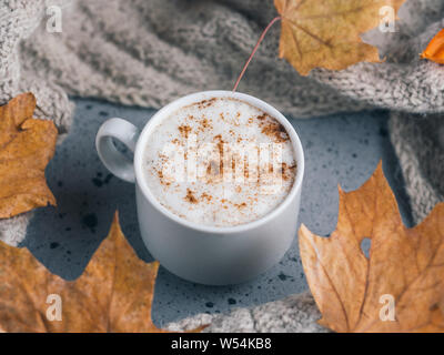 Weiße Tasse Latte Kaffee auf graue Tabelle mit einem gestrickten Schal und einem trockenen Herbst Ahorn Blätter. Home Atmosphäre und Komfort bei kalter Witterung Stockfoto