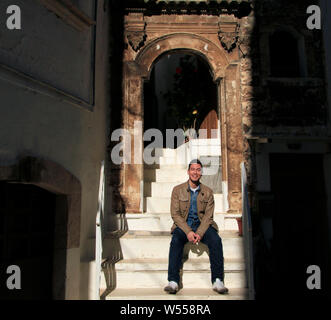 Lächelnden jungen Mann sitzt in der Sonne auf einer Treppe zu einem alten Gebäude in Italien. Stockfoto