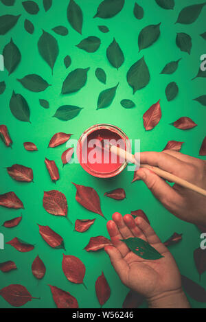 Malerei, grüne Blätter mit roter Farbe aus der Dose. Des Menschen Hand malen mit einem Bleistift die Blätter rot Stockfoto