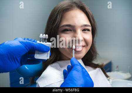 Zahnarzt in Blau medizinische Handschuhe, die Probe, die von Zahnschmelz Maßstab zu lächelnde Frau Patienten bis rechten Farbton für Zähne bleichen Verfahren auswählen Stockfoto