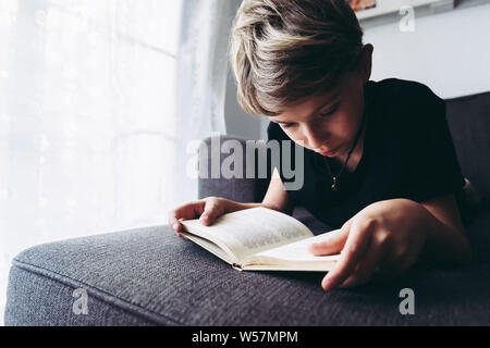 Nahaufnahme der blonde Junge lesen Buch konzentriert Student liegend liest die Lektion sehr entspannt, sorgfältige Kind liest eine Geschichte, junger Mann Stockfoto