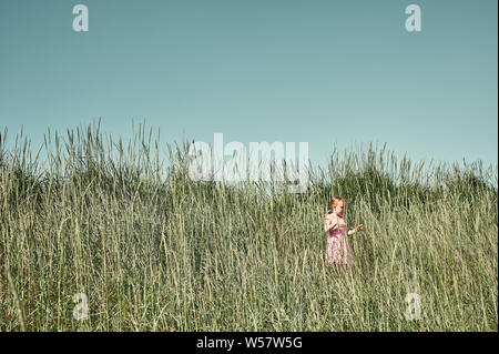 Hübsche junge Mädchen in rosa Kleid wandern im hohen Gras in der üppigen sonnendurchfluteten grünen Feld auf Grotta Insel im Sommer Stockfoto