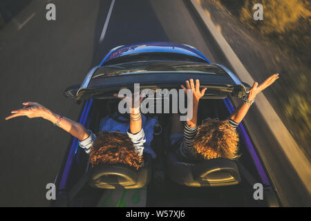 Ansicht von oben ein Paar euphorischen Frauen in einem Cabrio verdrehen und winkend. Zwei lockiges Mädchen im Urlaub Spaß fahren Auto, glücklich lachend Stockfoto