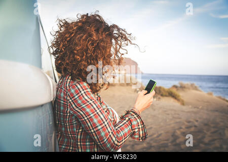 Nicht erkennbare Mädchen mit handy Seashore, lehnte sich gegen Vintage van, Auburn lockiges Haar zerzauste Wind, Frau mittleren Alters Ferienhäuser Insel chatten Stockfoto