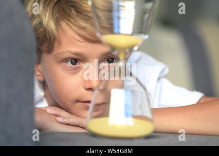 Schön blond lächelnd Kerl liegt mit seinen Händen unter sein Gesicht schaut verträumt fasziniert sich langsam bewegenden Sand in Glas Sanduhr. gespannter Blick, Konzept von de Stockfoto