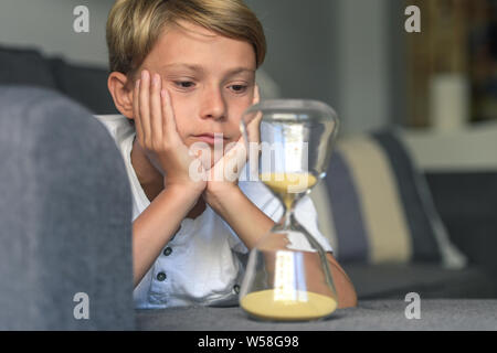 Schön blond lächelnd Kerl liegt mit seinen Händen unter sein Gesicht schaut verträumt fasziniert sich langsam bewegenden Sand in Glas Sanduhr. gespannter Blick, Konzept von de Stockfoto