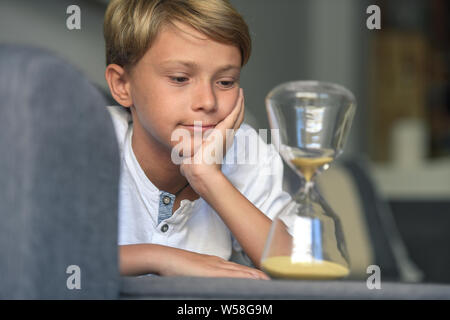 Schön blond lächelnd Kerl liegt mit seinen Händen unter sein Gesicht schaut verträumt fasziniert sich langsam bewegenden Sand in Glas Sanduhr. gespannter Blick, Konzept von de Stockfoto