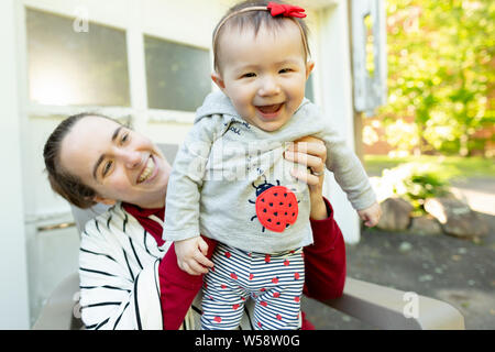 Baby Mädchen steht auf Mutters Schoß lächelnd, während Mutter liebevoll schaut Stockfoto