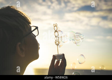 Junge blonde Junge bläst eine lange Reihe von Blasen in ein Feld; niedliche Kind vor dem Meer jagen Seife kochen, schönes Kind spielen im Freien in einem schönen Stockfoto