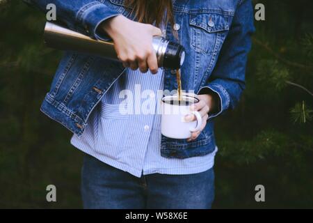 Reisende Mädchen gießt Kaffee aus der Thermoskanne Schale, im Freien. Junge Frau trinkt Kaffee in der Tasse. Thema Reisen. Frau gießen ein heißes Getränk in Becher aus der Thermoskanne. Gi Stockfoto