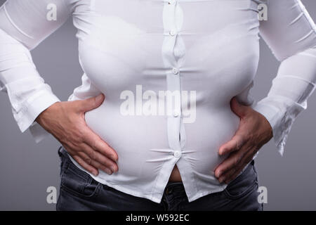 Den mittleren Abschnitt der Fette Frau mit Hemd zu klein berühren Ihren Bauch gegen grauer Hintergrund Stockfoto