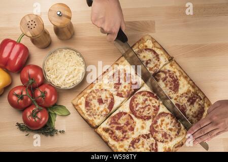 Ein Schuss von einer Person, die eine quadratische Pepperoni-Pizza schneidet Mit Gemüse auf der Seite Stockfoto