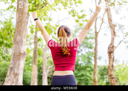 Junge Frau wirft beide Arme in fröhlichen Feier nach Ihrem Morgen übung Finishing an einem Park, unscharfen Bäume und Himmel Hintergrund, positive Er Stockfoto