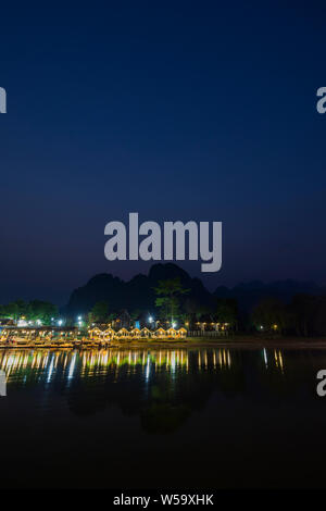 Wenige Leute sitzen an einem beleuchteten Restaurant Waterfront durch den Nam Song Fluss und die Silhouette von Karst Kalkstein Berge in Vang Vieng, Laos, in der Dämmerung. Stockfoto