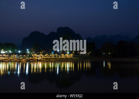 Wenige Leute sitzen an einem beleuchteten Restaurant Waterfront durch den Nam Song Fluss und die Silhouette von Karst Kalkstein Berge in Vang Vieng, Laos, in der Dämmerung. Stockfoto