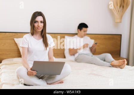 Portrait von Mädchen Arbeiten am Laptop im Bett. Kerl in Phone hinter seiner Freundin. Junge Frau mit Kamera schaut. Mädchen ist die Eingabe auf Editor, der Kerl ist Stockfoto