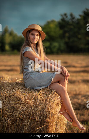 Schönes junges Mädchen mit langen blonden Haaren in einen Strohhut sitzend auf einem Ballen Stroh. Sommer Sonnenuntergang. Stockfoto