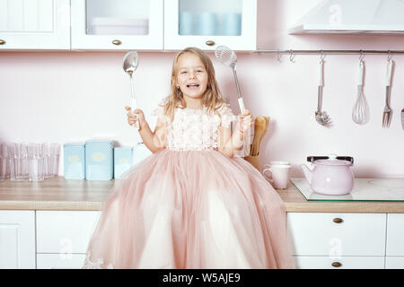 Wunderschöne kleine Mädchen, das in einem prächtigen Kleid sitzt auf dem Tisch in der Küche und lacht. Stockfoto