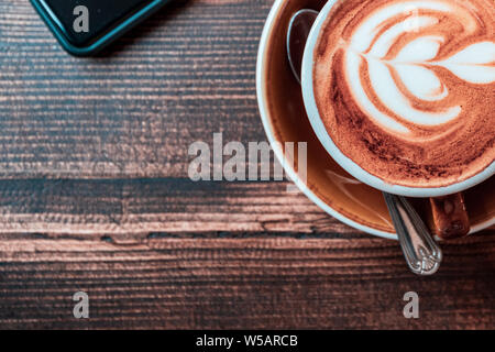 Ein Schuß von oben auf ein Telefon und eine Tasse Cappuccino auf einem dunklen Holztisch körnig Stockfoto