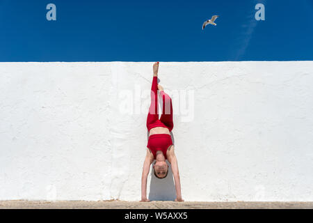 Frau macht Handstand gegen eine weiße Wand mit einem blauen Himmel tragen rot. Stockfoto