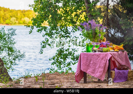 Picknick in der Nähe Wasser am Tisch mit Blumen im Wald Obst und Gemüse Stockfoto