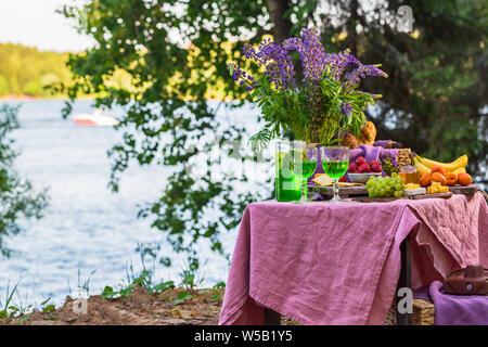 Picknick in der Nähe Wasser am Tisch mit Blumen im Wald Obst und Gemüse Stockfoto