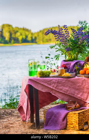 Picknick in der Nähe Wasser am Tisch mit Blumen im Wald Obst und Gemüse Stockfoto