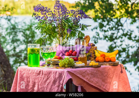 Picknick in der Nähe Wasser am Tisch mit Blumen im Wald Obst und Gemüse Stockfoto