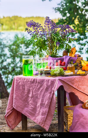 Picknick in der Nähe Wasser am Tisch mit Blumen im Wald Obst und Gemüse Stockfoto