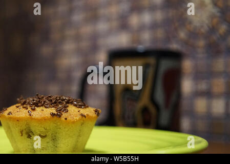 Cupcake mit Chocolate Chips auf einem grünen Untertasse close-up. Im Hintergrund eine schwarze Tasse Tee und Mosaik Wand Stockfoto